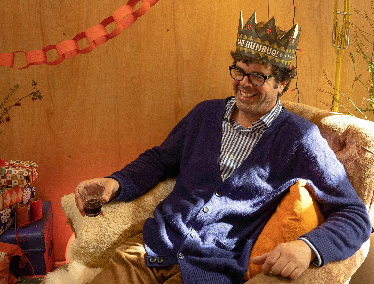 Man sitting in a chair wearing a party hat that says bah humbug on it with streamers and a lamp in the background.
